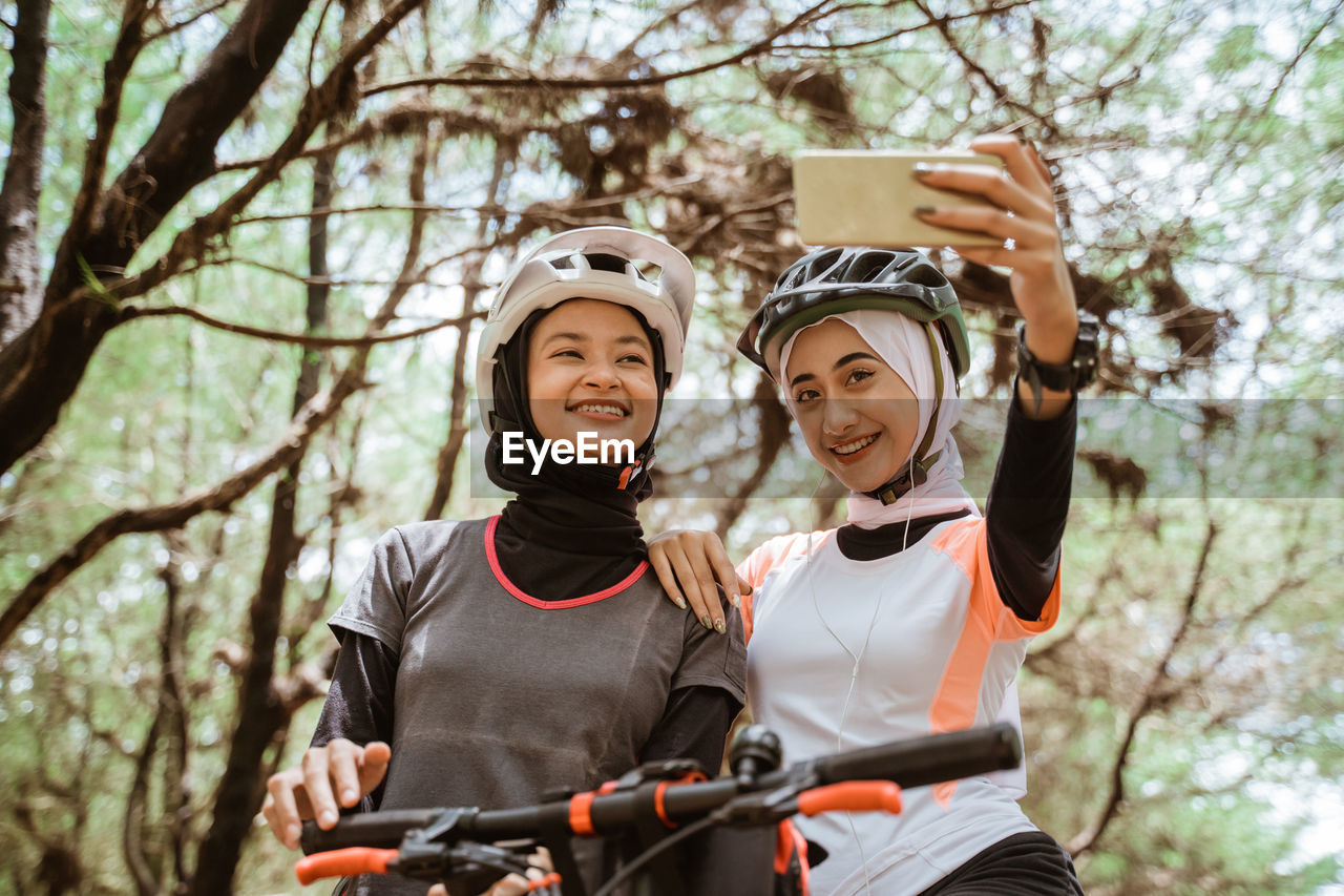 Smiling young females taking selfie against trees