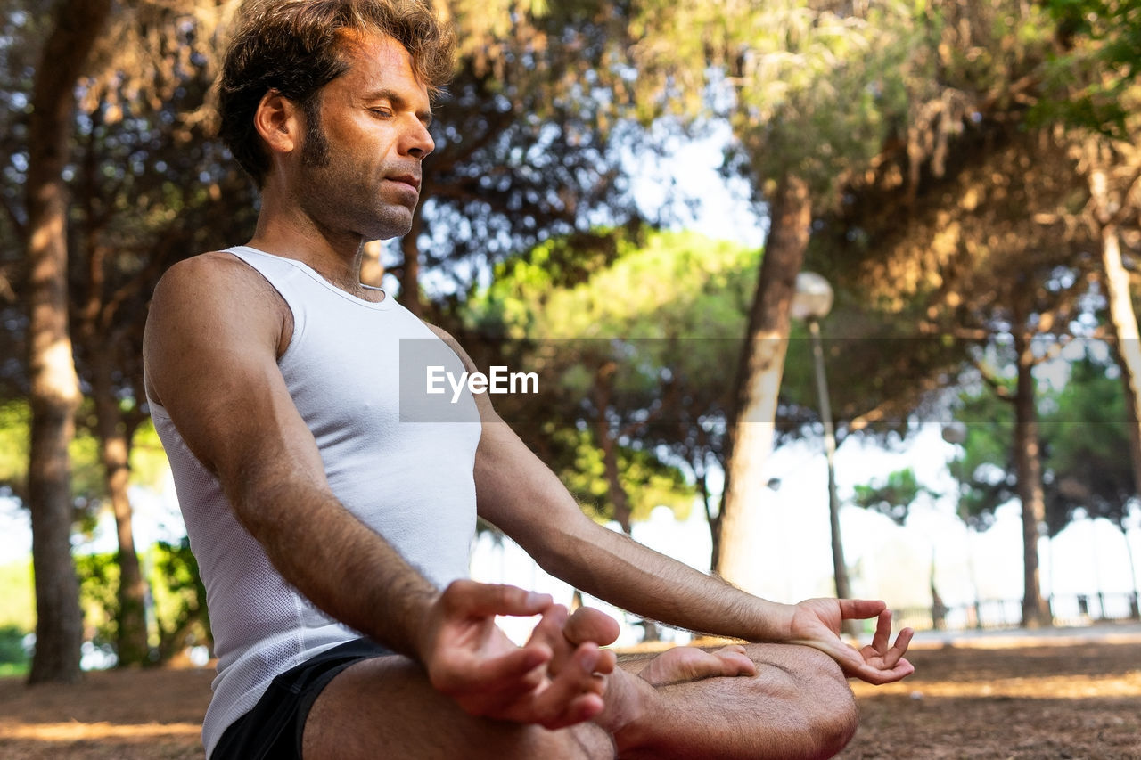 From below side view of barefoot focused male sitting in lotus pose with eyes closed on magenta mat and gesturing mudra during meditation session on sunny summer day in park