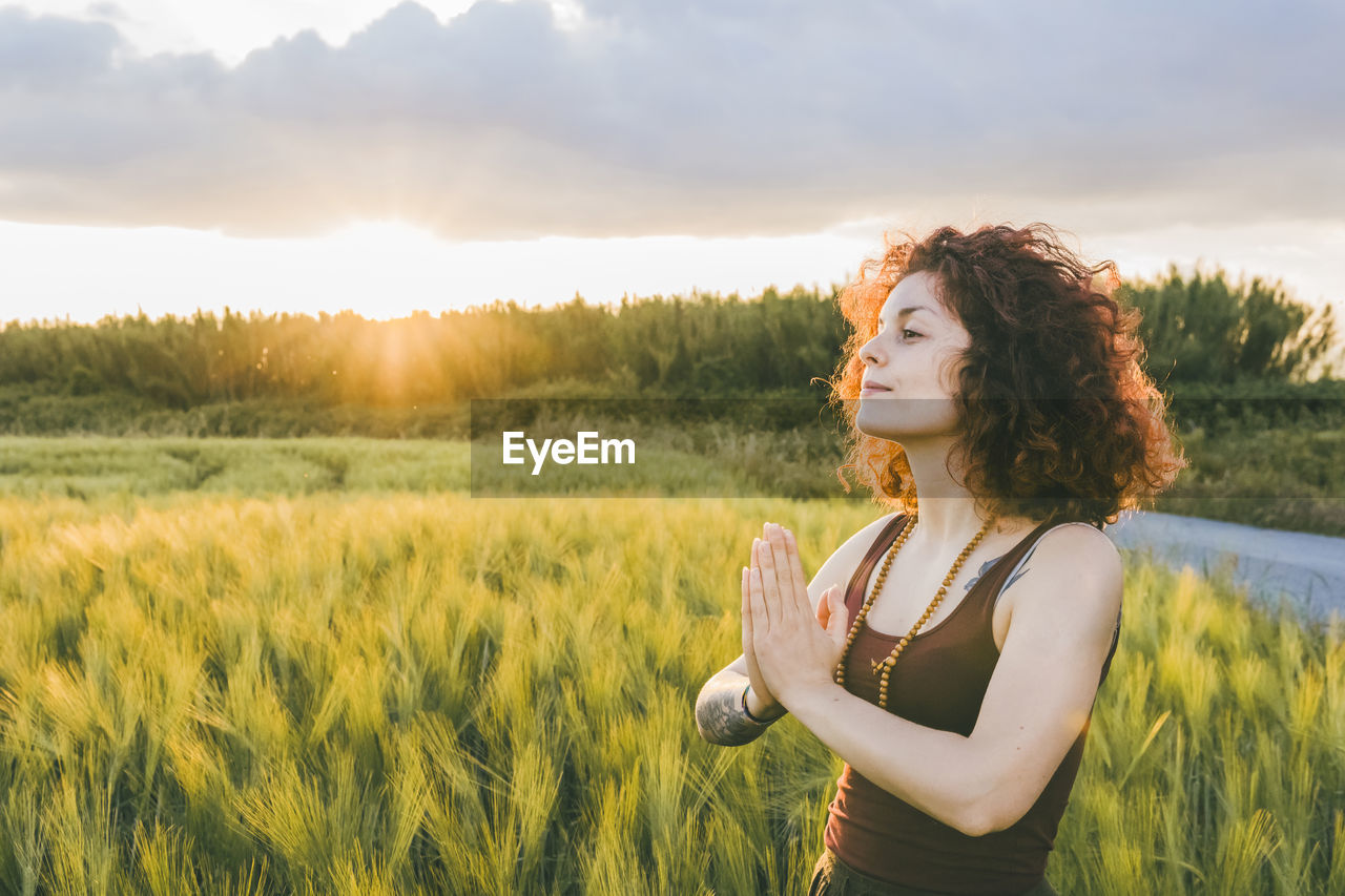 Woman meditating while standing at farm against cloudy sky during sunset