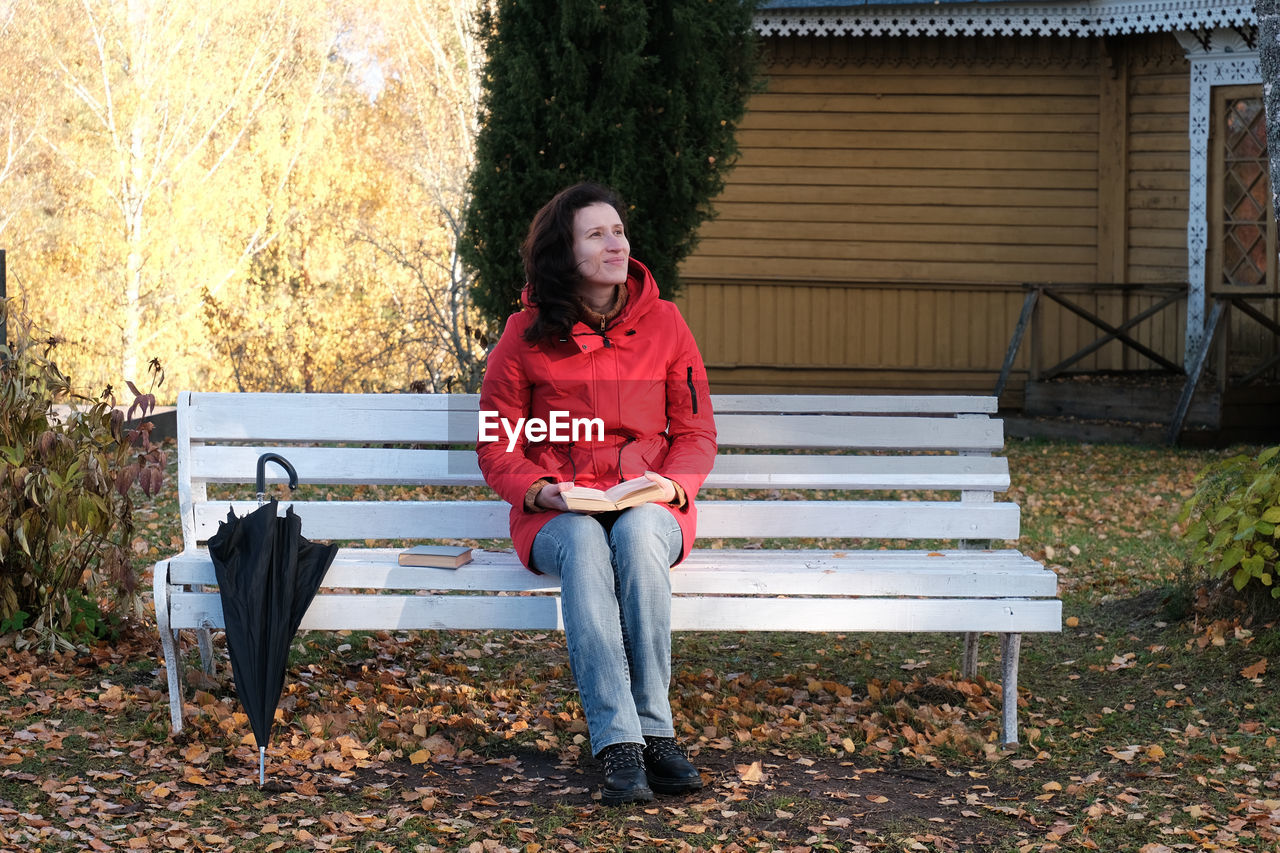 A woman in a red jacket is sitting on a white bench in the park and reading a paper book.