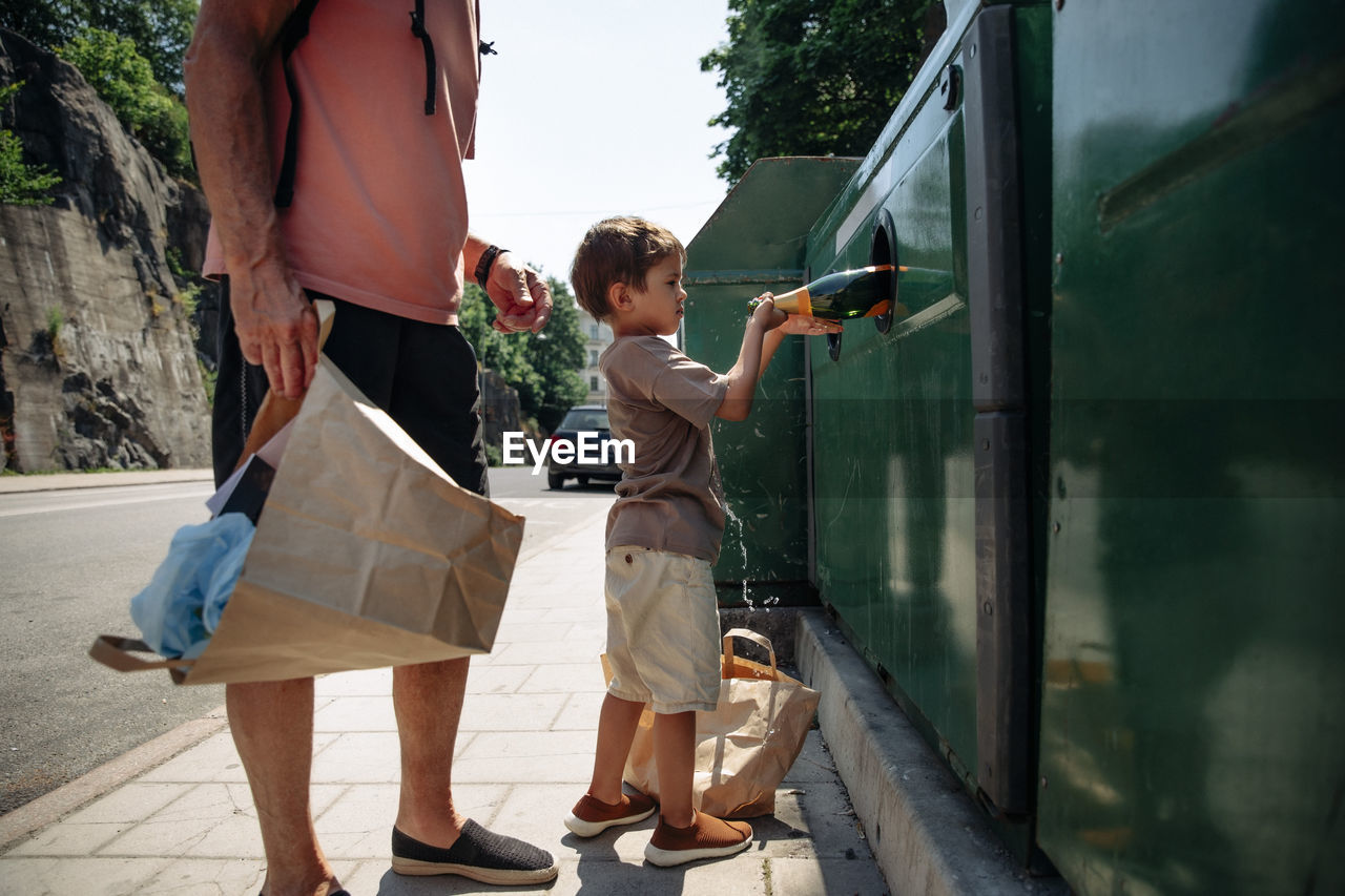 Side view of boy trashing bottle in garbage bin while standing with grandfather
