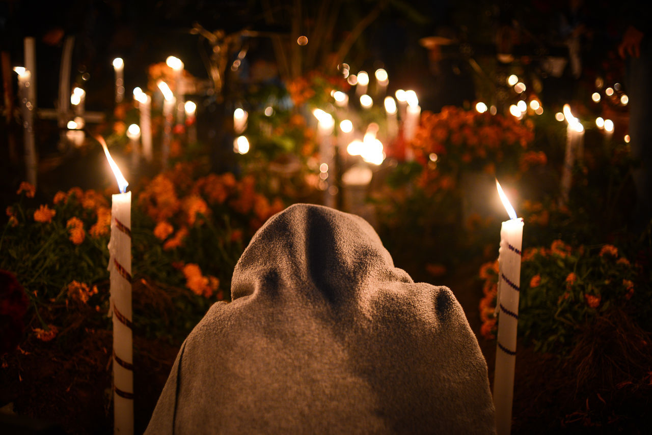 Rear view of man covered with blanket walking on lit cemetery at night