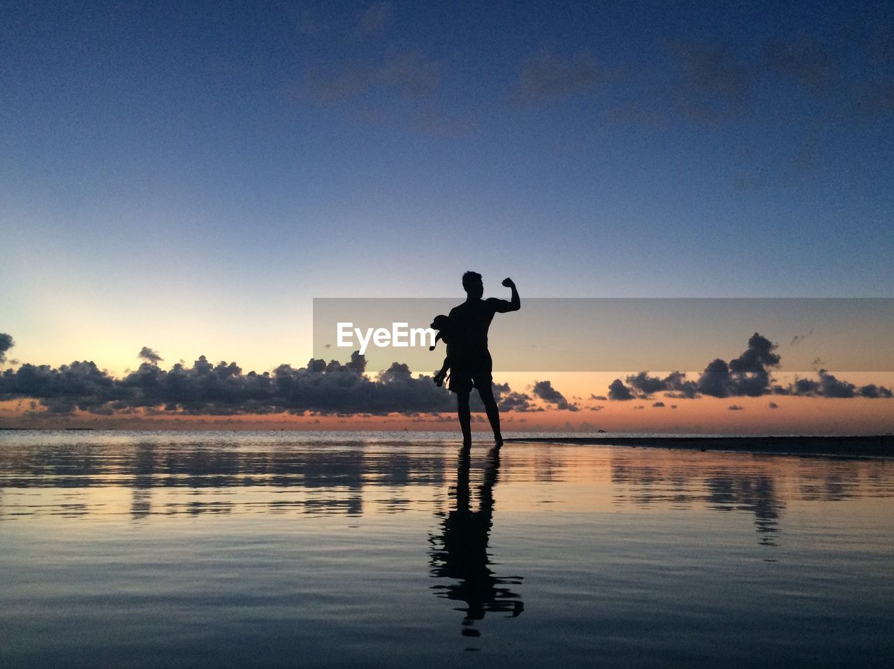 Silhouette father flexing muscles while carrying child at beach during sunset