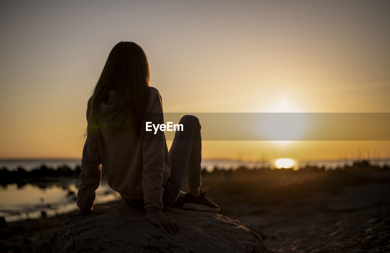 Rear view of girl sitting at beach during sunset