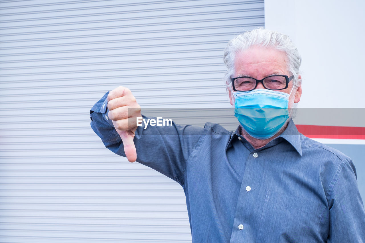 Portrait of man wearing mask gesturing thumb down while standing against shutter