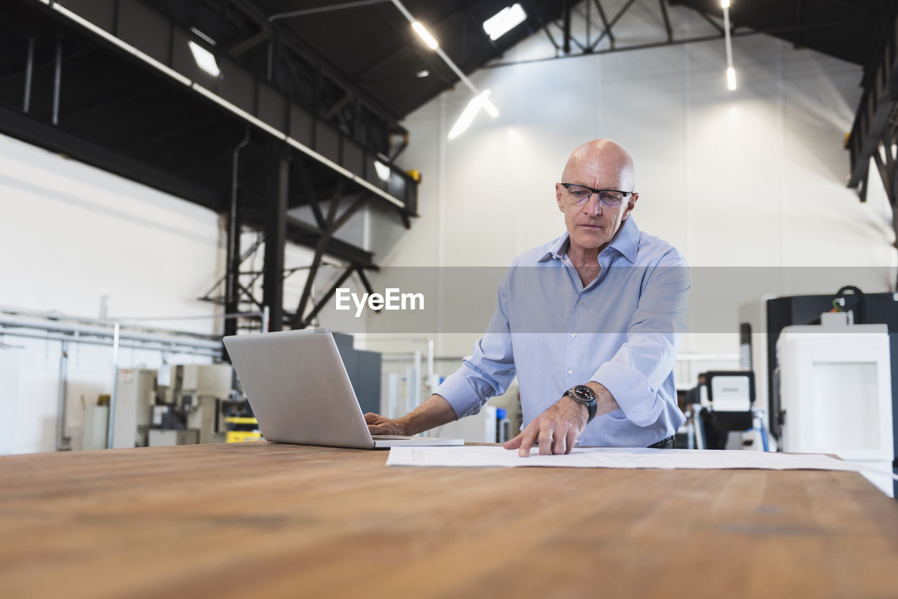 Man with laptop looking at plan on table in factory