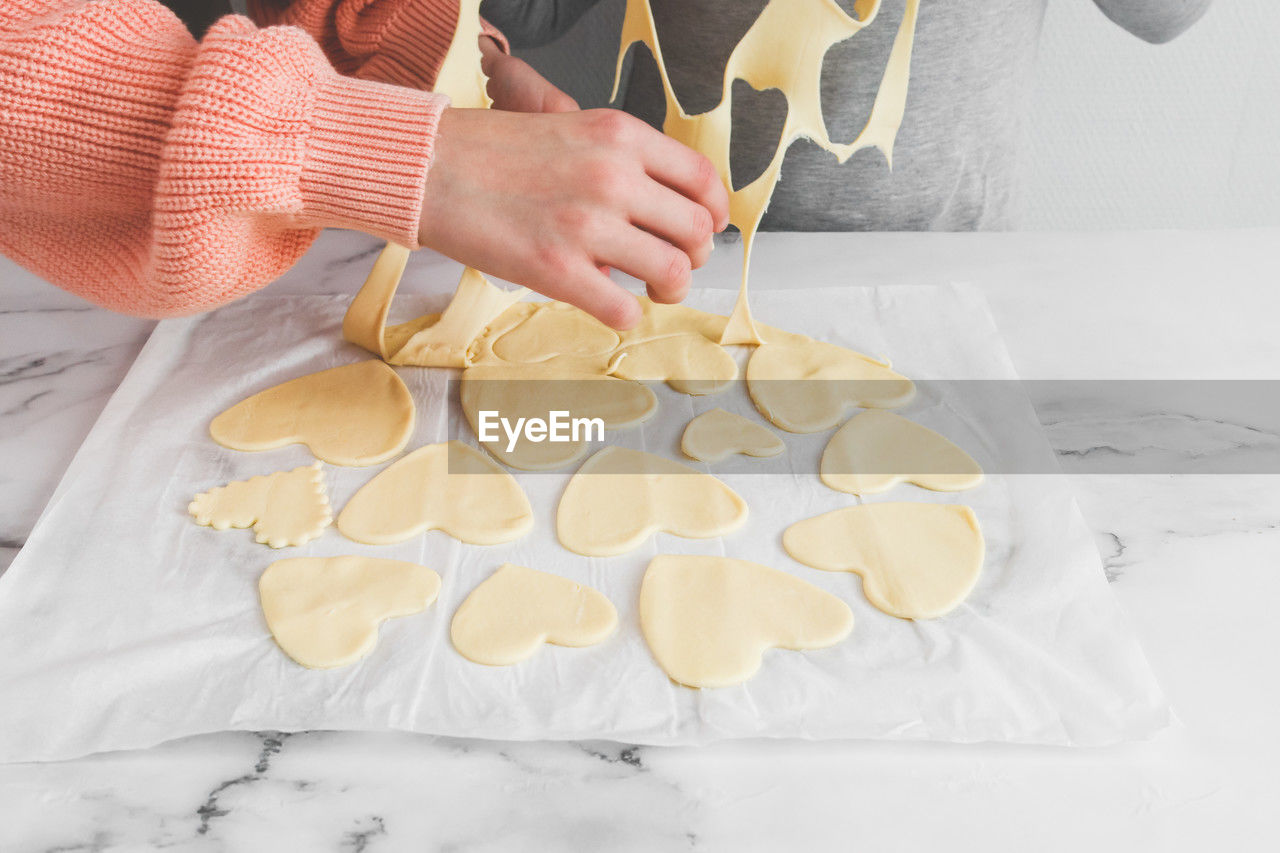 Hands of a young caucasian girl of a baker's sister separating raw dough from cut out hearts.