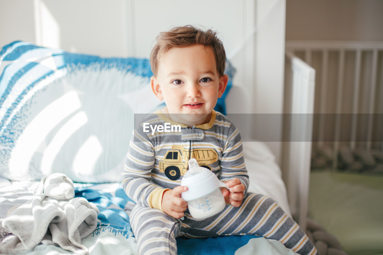 Cute adorable caucasian kid boy sitting on bed drinking milk from kids bottle. healthy eating 