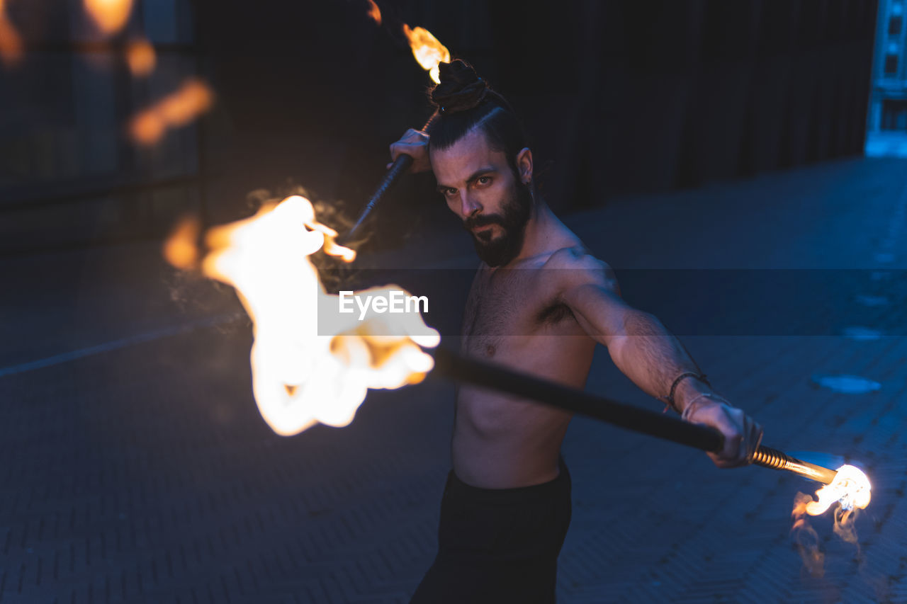 Hipster male juggling with fire staff on footpath at dusk
