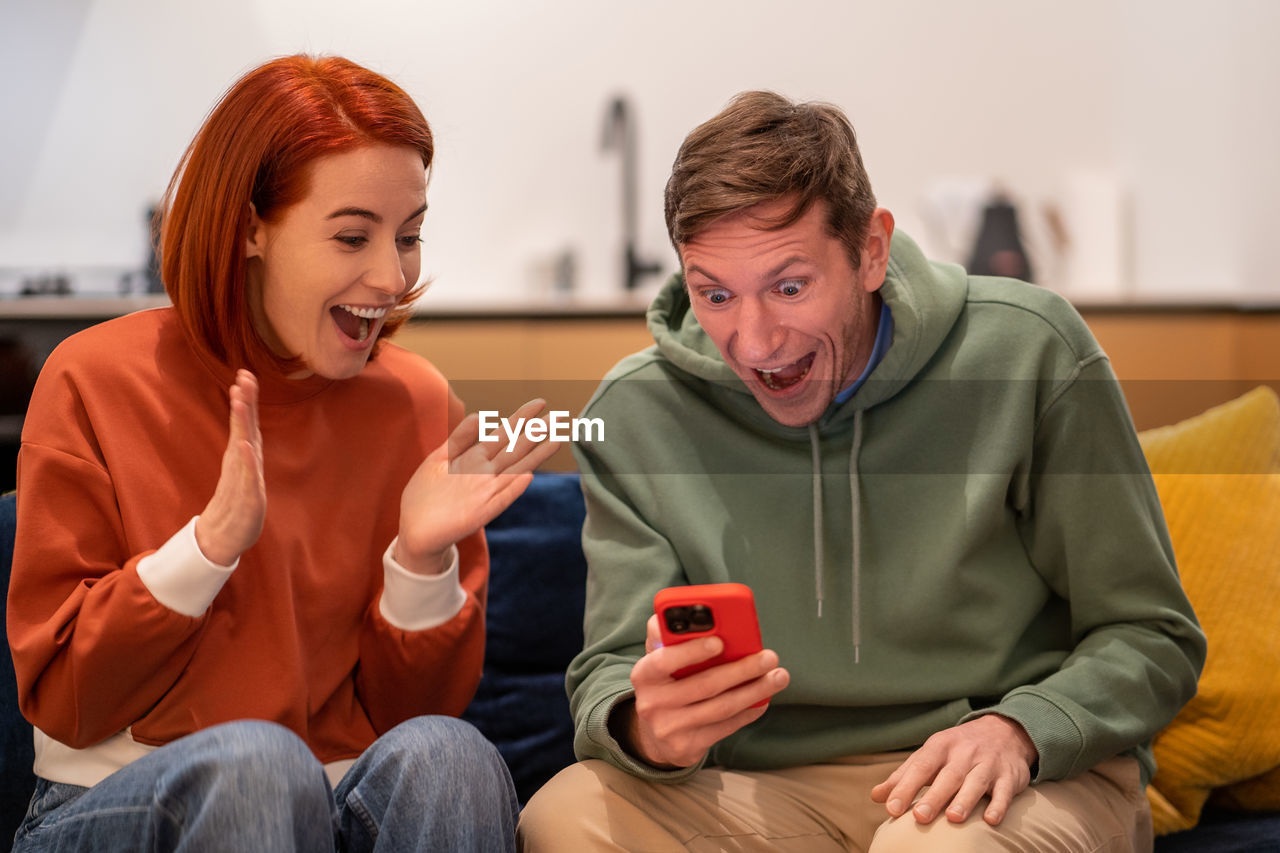 Excited overjoyed millennial happy young family couple looking at phone screen screaming with joy