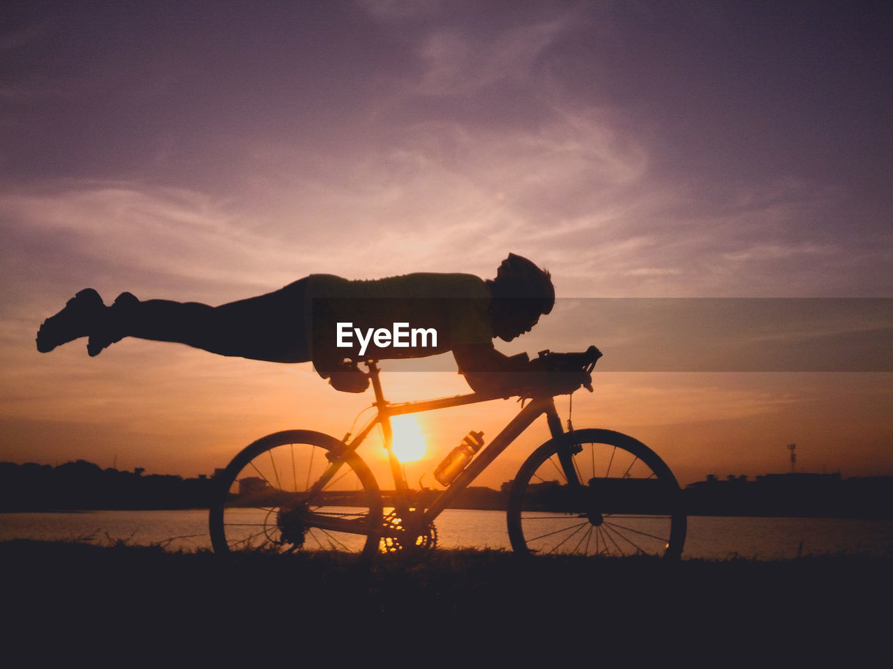 Silhouette man balancing on bicycle seat by lake against sky during sunset