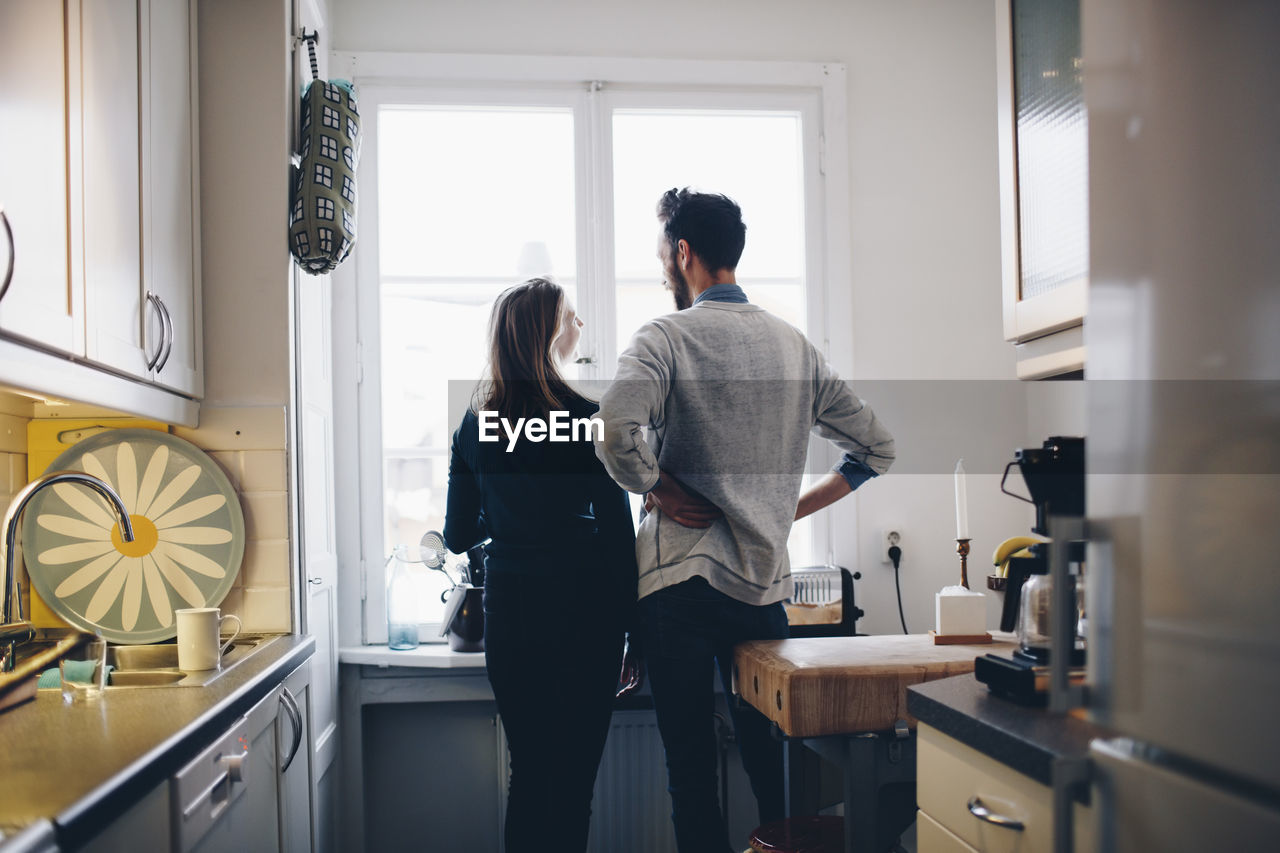 Rear view of couple talking while standing by dining table against window at home