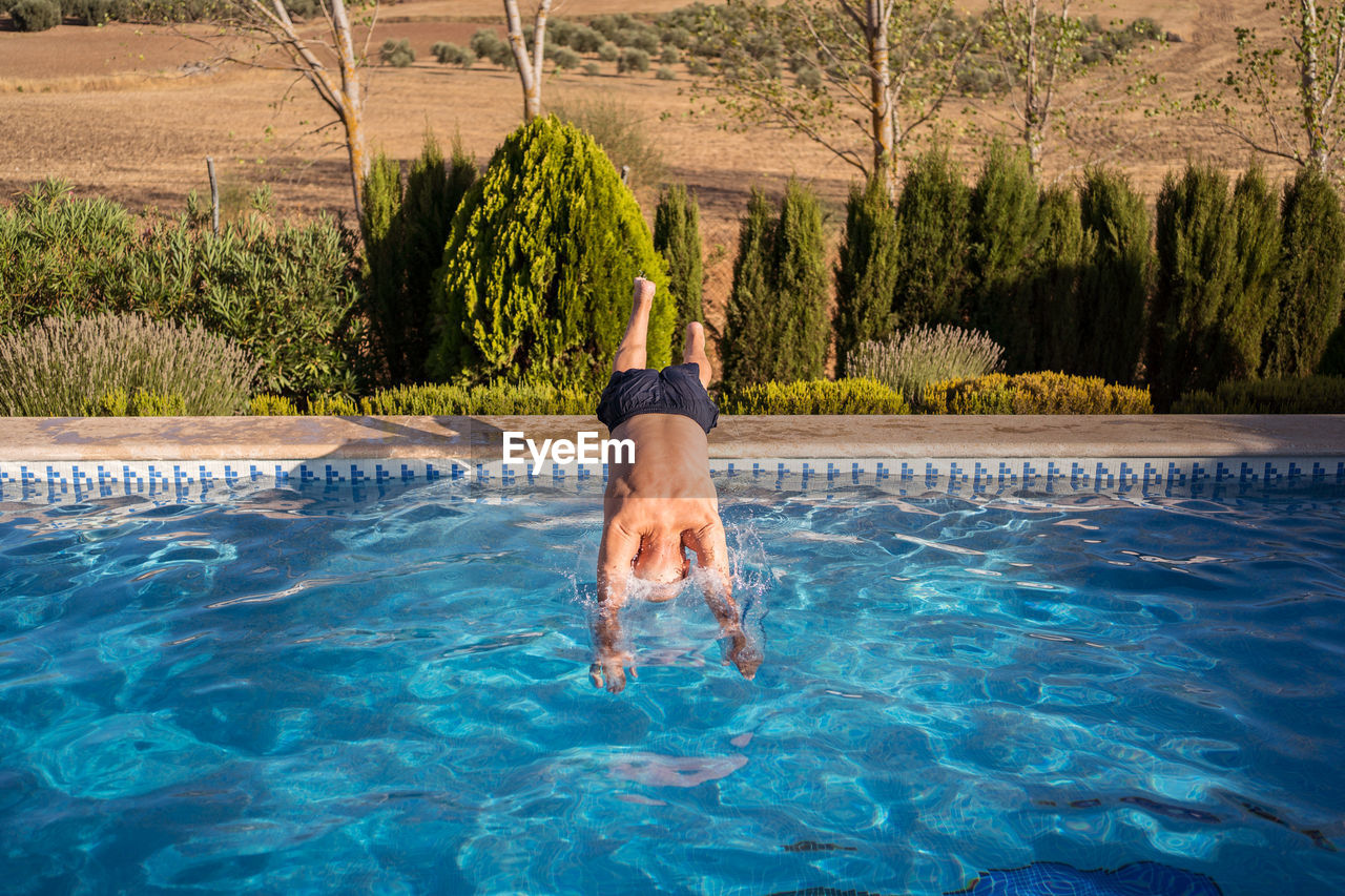 Full body back view of unrecognizable barefoot and shirtless senior male jumping in swimming pool with clear blue water