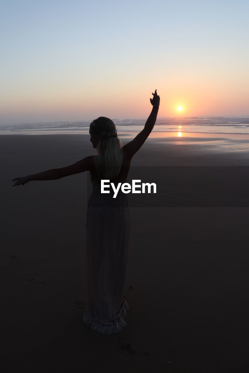 REAR VIEW OF WOMAN STANDING ON BEACH AGAINST SKY DURING SUNSET