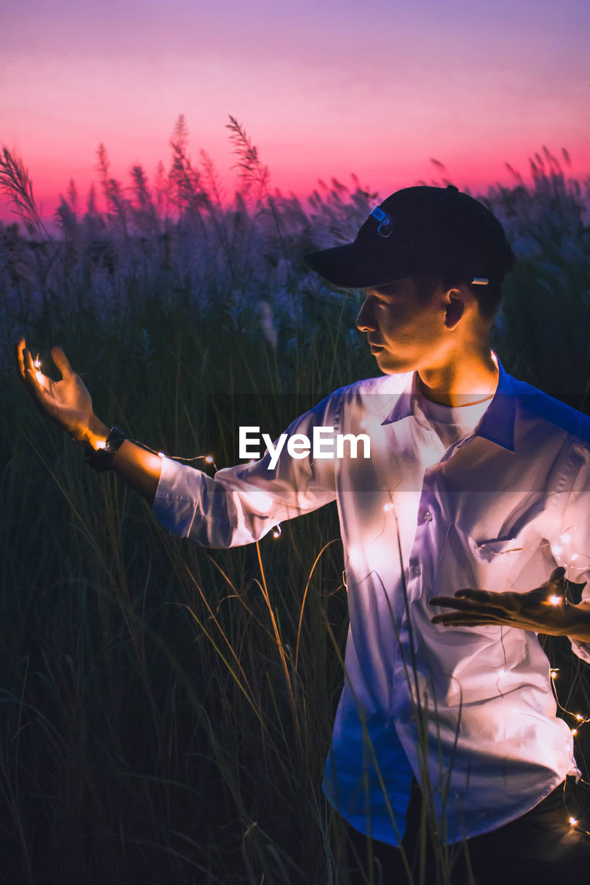 Young man holding illuminated string light while standing on field during dusk