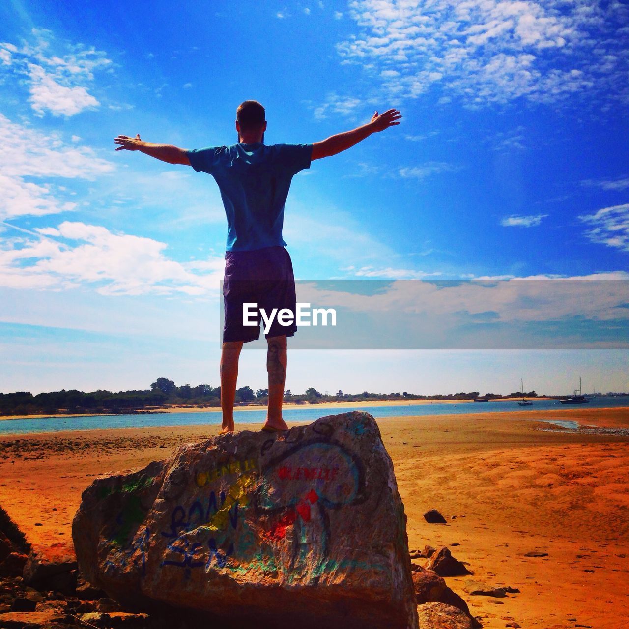 Rear view of man with arms outstretched on rock at beach against sky