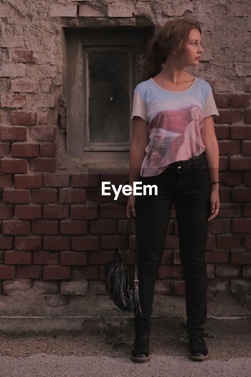 Full length of thoughtful young woman standing against brick wall