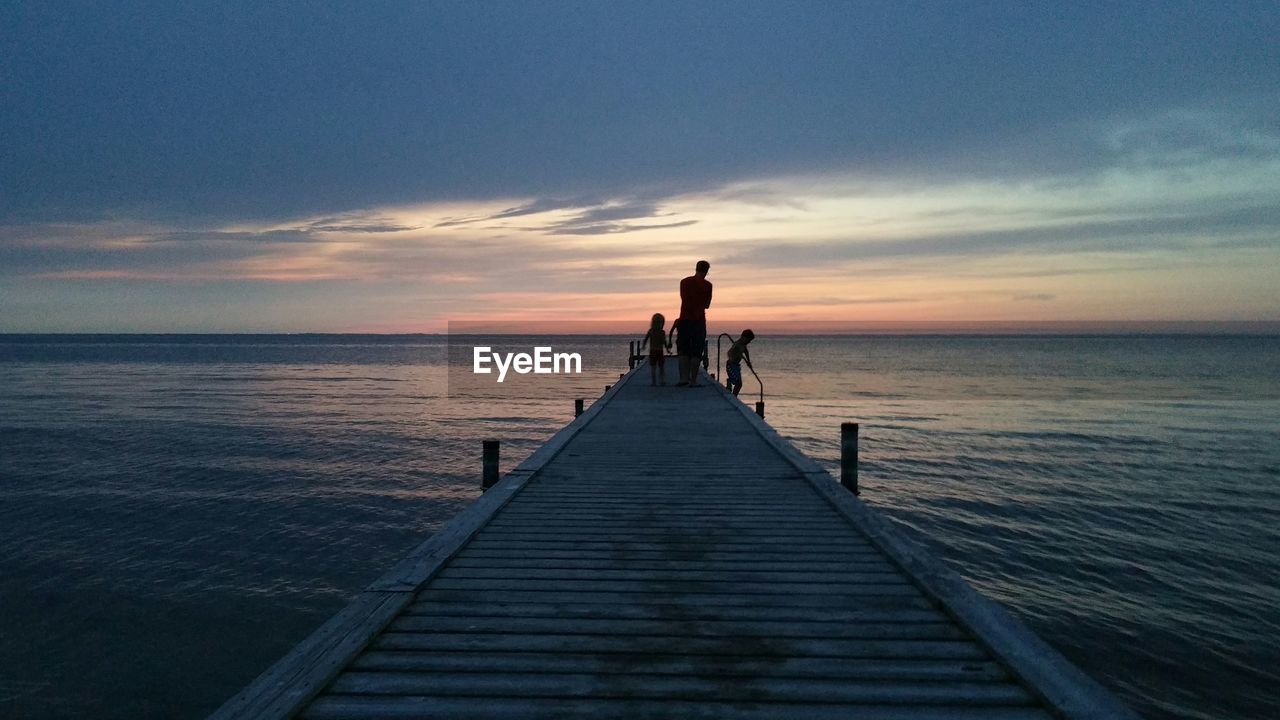 Silhouette father with children on pier over sea against cloudy sky during sunset