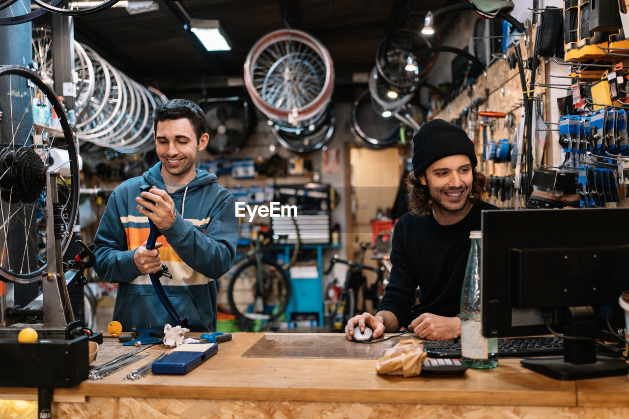 Positive young male technician assembling bike while colleague working on computer in professional workshop