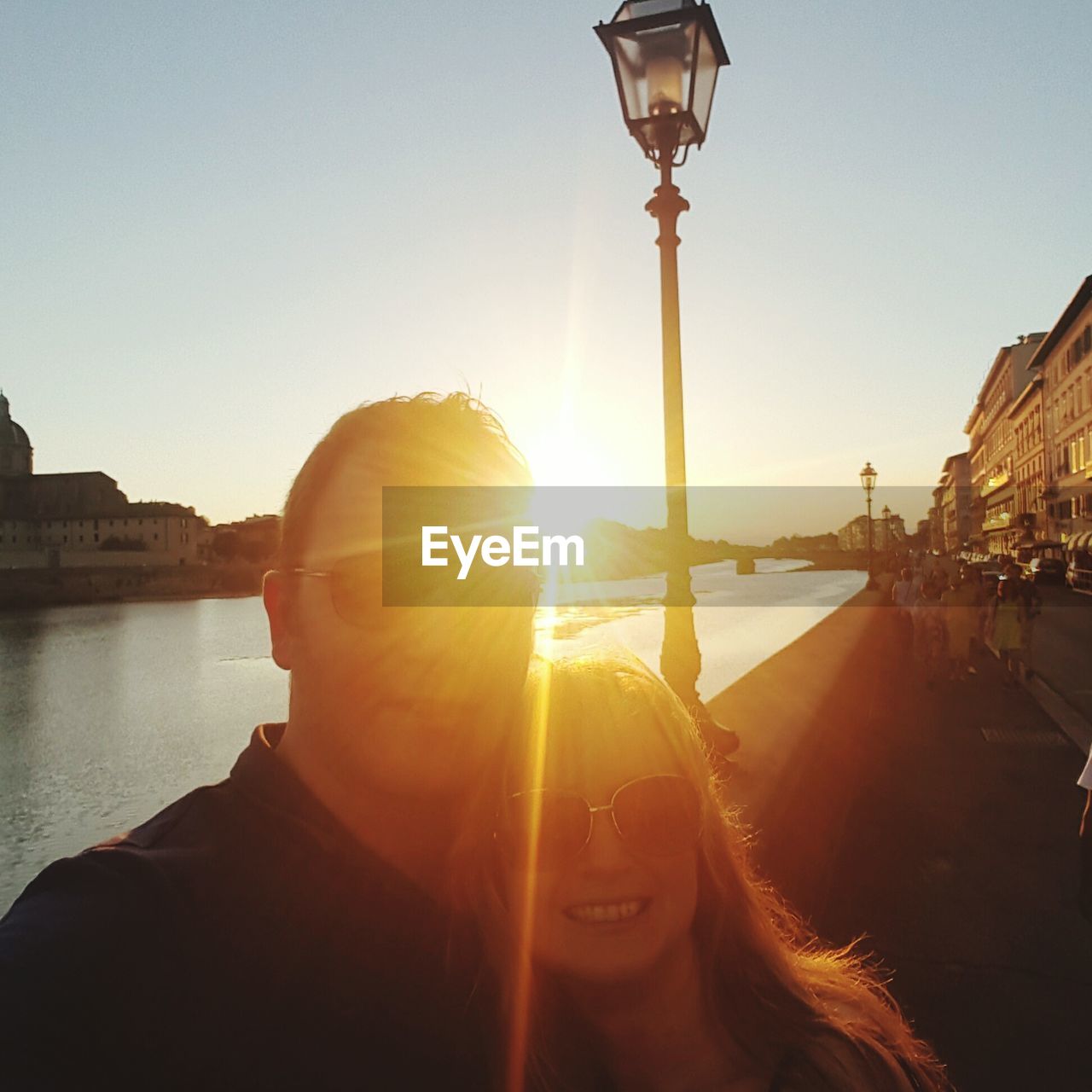 Couple standing on sidewalk by river in city on sunny day