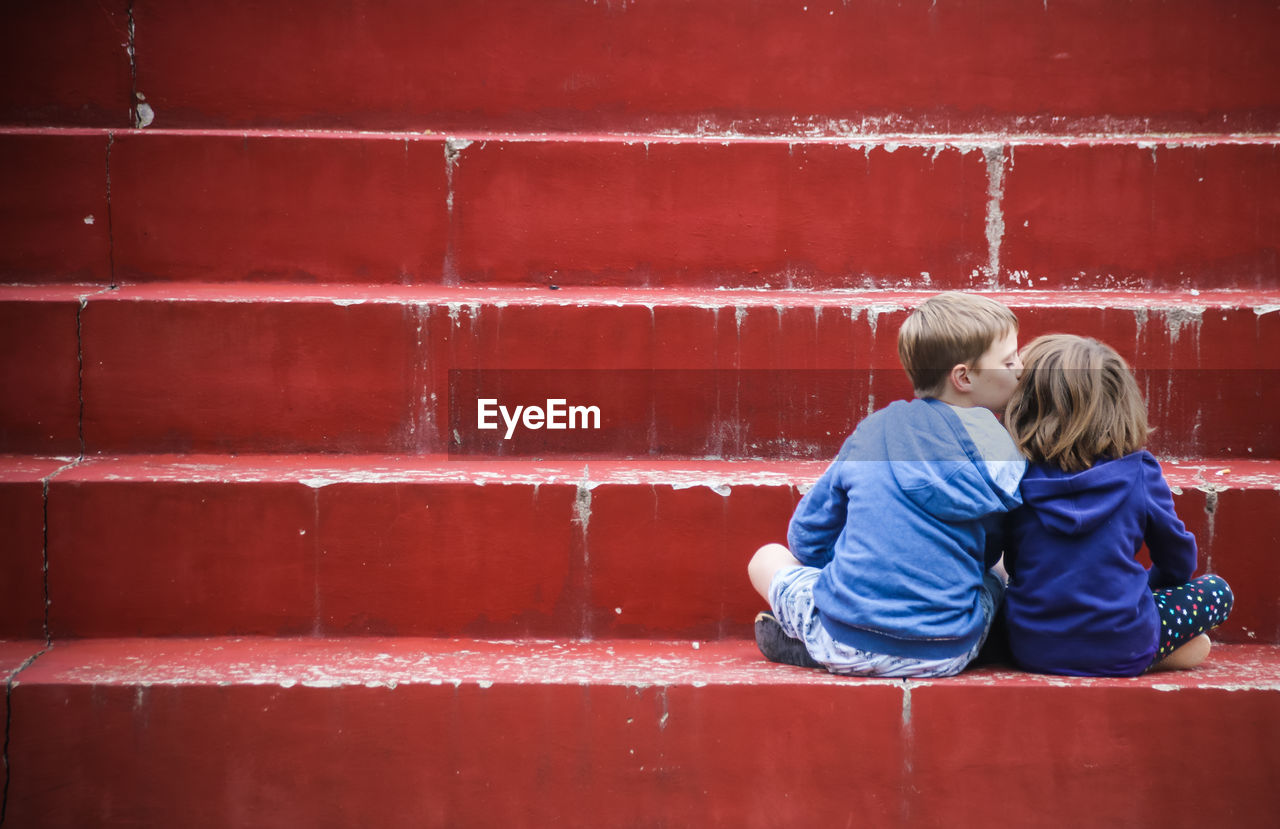 Rear view of boy kissing a girl and sitting against red wall
