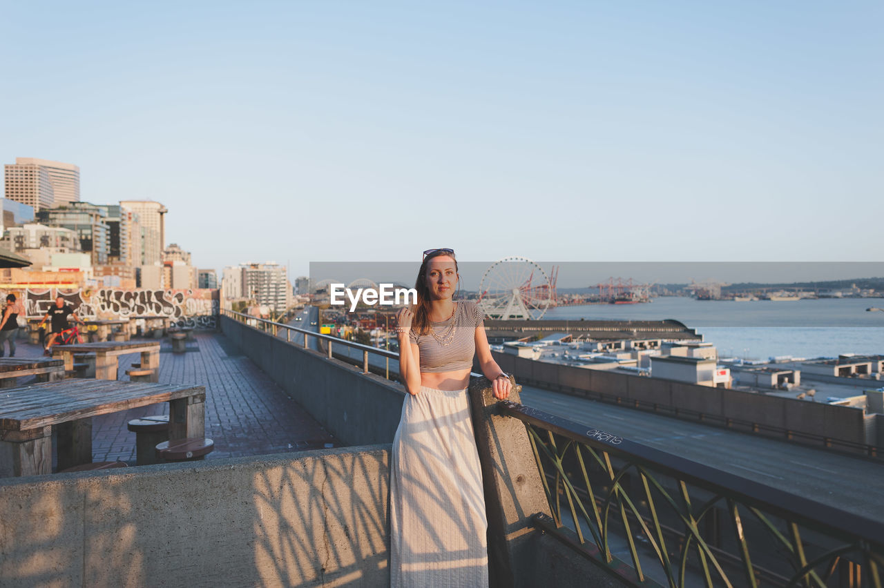 Portrait of beautiful woman standing on building terrace in city against clear sky