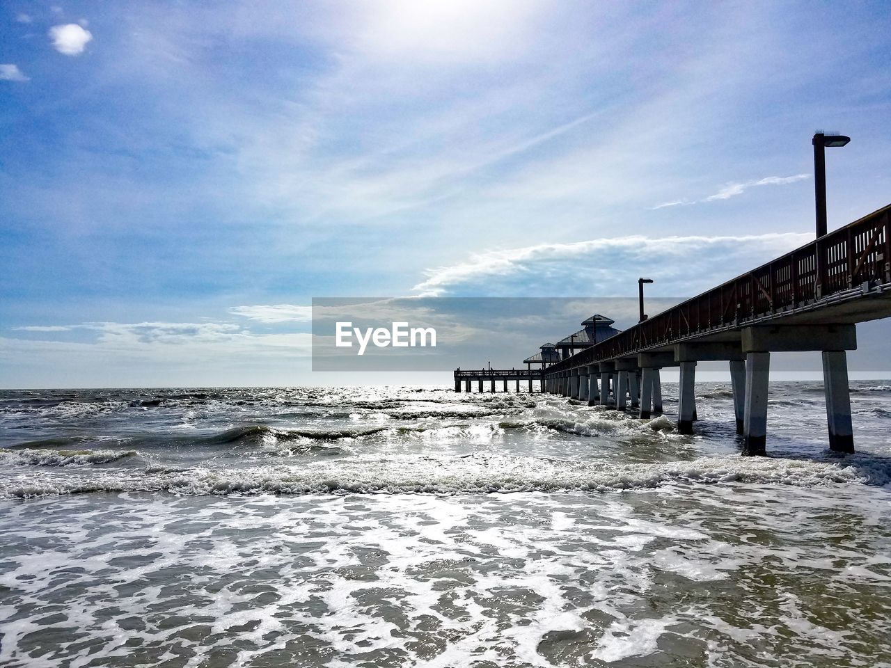 Scenic ocean view of breaking waves against a sunny sky with clouds and a long pier