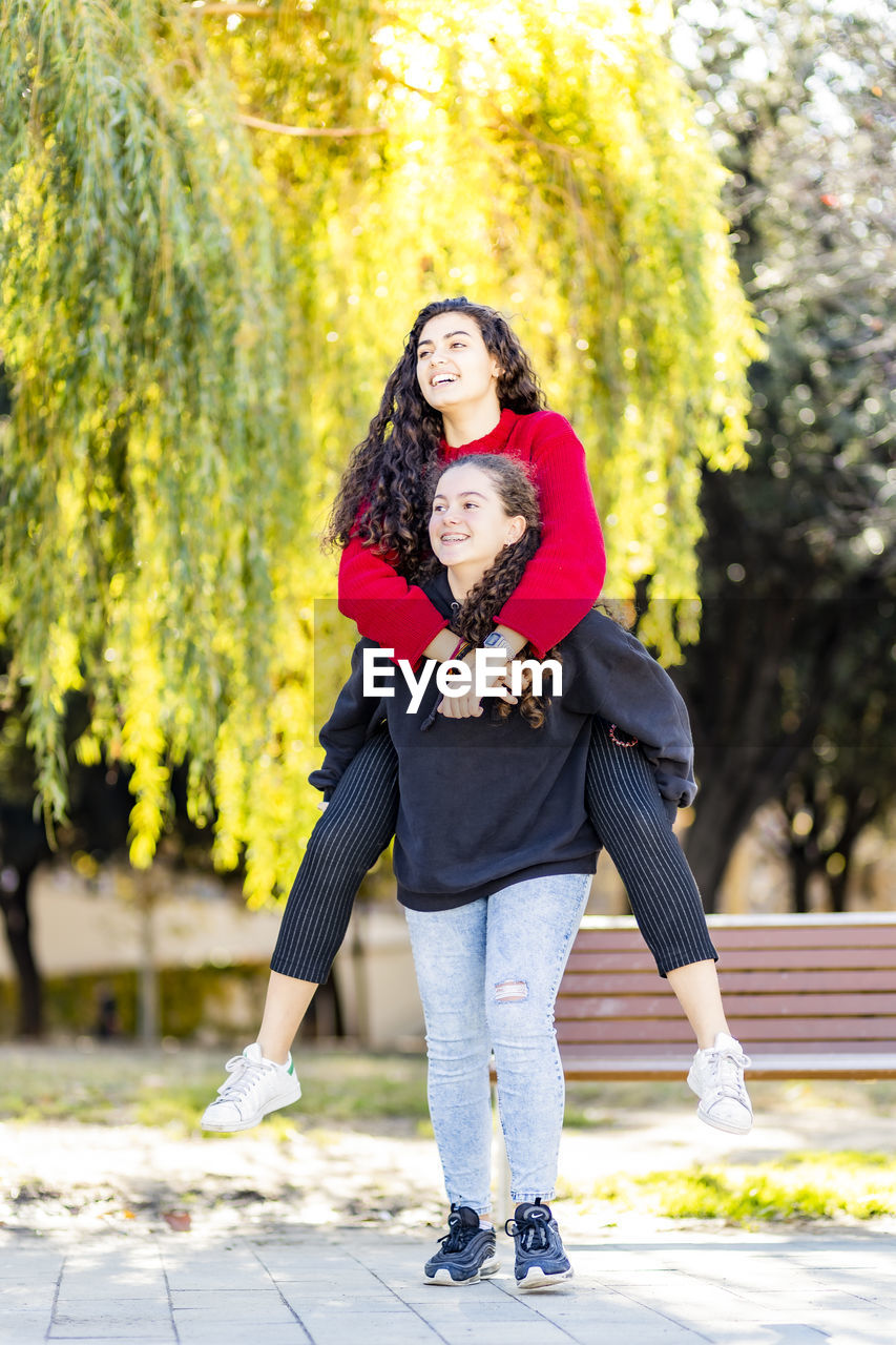 Happy teenage girl giving piggyback ride to friend in park