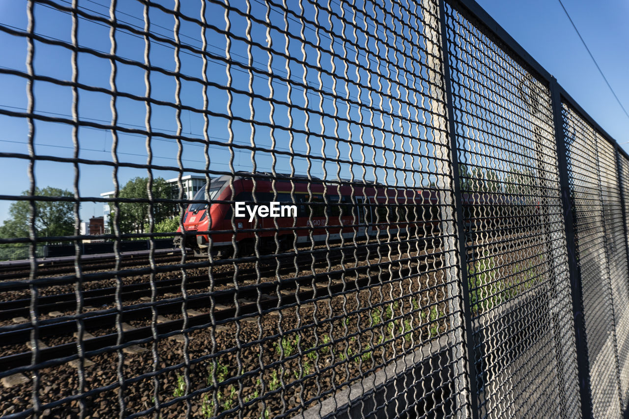 Train on railroad track seen through metallic fence against clear sky