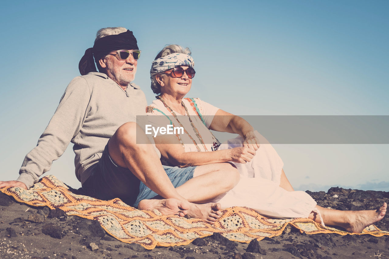Senior couple wearing sunglasses while sitting at beach against clear sky