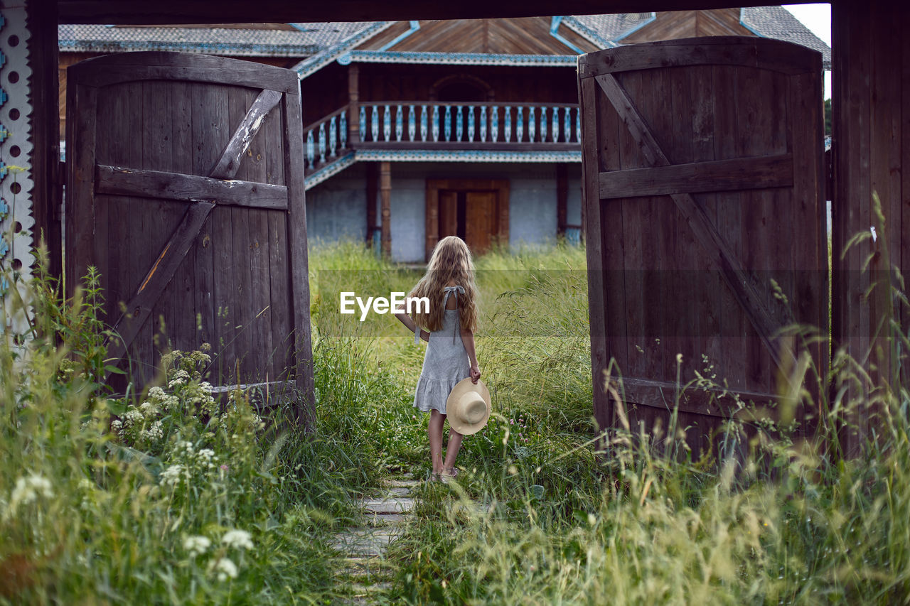 Blonde village girl with long hair stands in a wooden gate in the village in summer