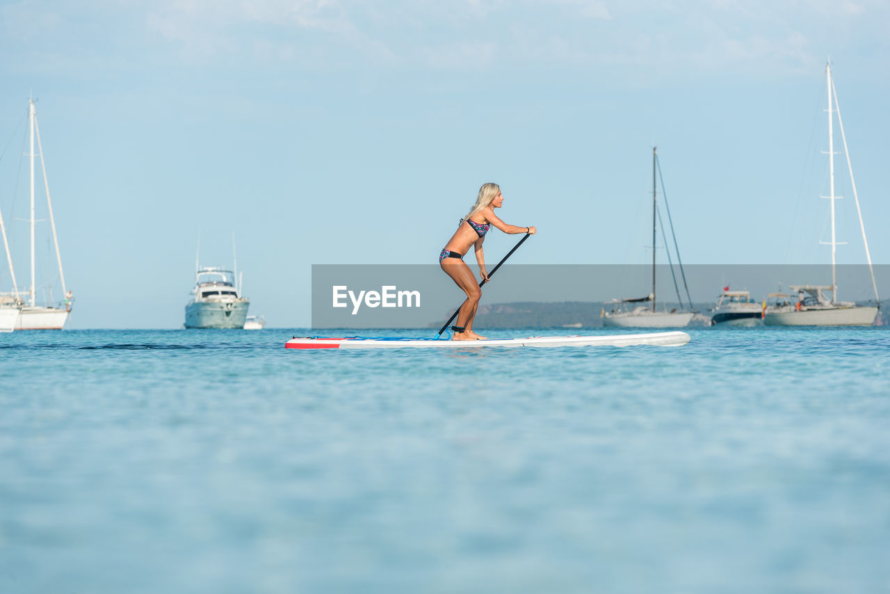 Side view of fit female surfer in bikini standing on paddleboard and rowing while training in sea on sunny day
