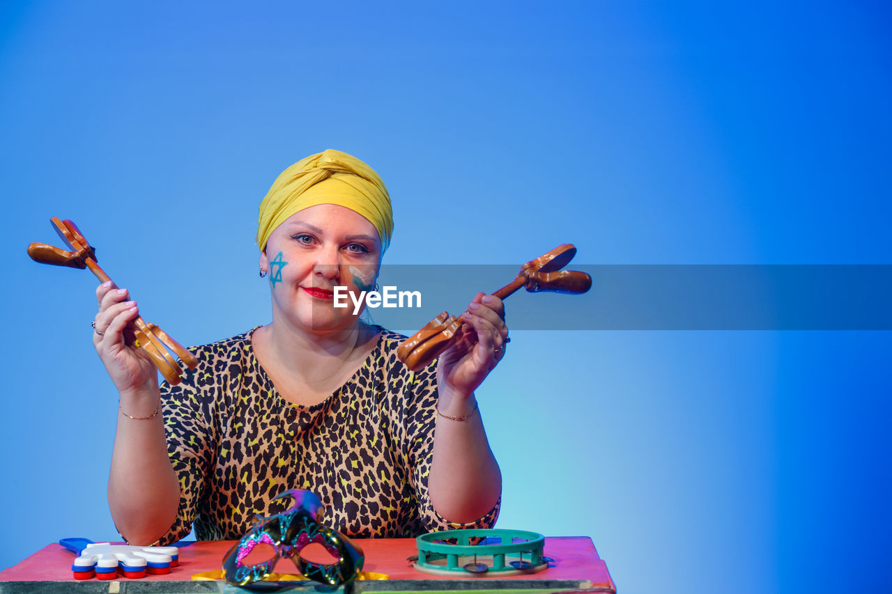 one person, portrait, blue, adult, smiling, women, happiness, emotion, looking at camera, front view, blue background, waist up, fun, copy space, arts culture and entertainment, colored background, toy, person, holding, indoors, studio shot, cheerful, female, young adult, enjoyment, occupation, fashion, multi colored