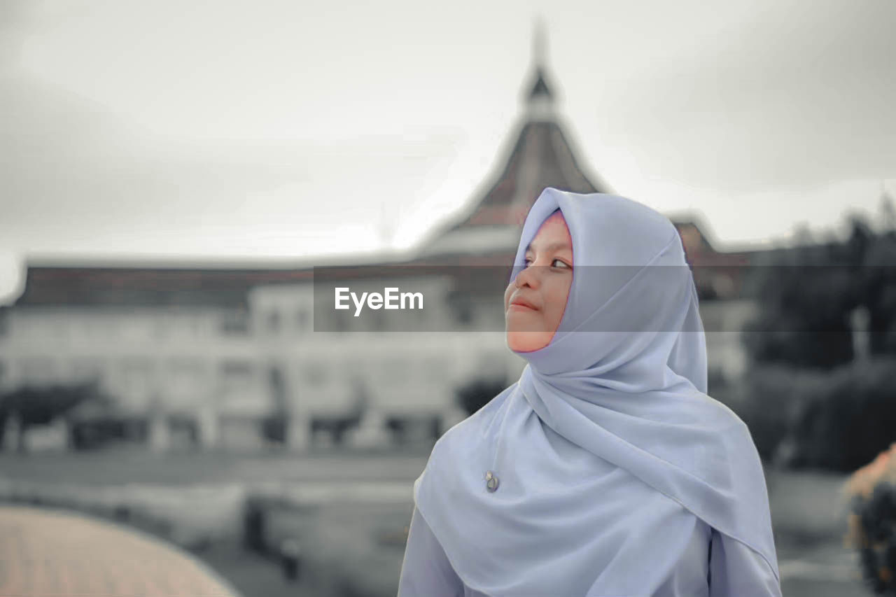 Young woman wearing hijab looking away against sky in city
