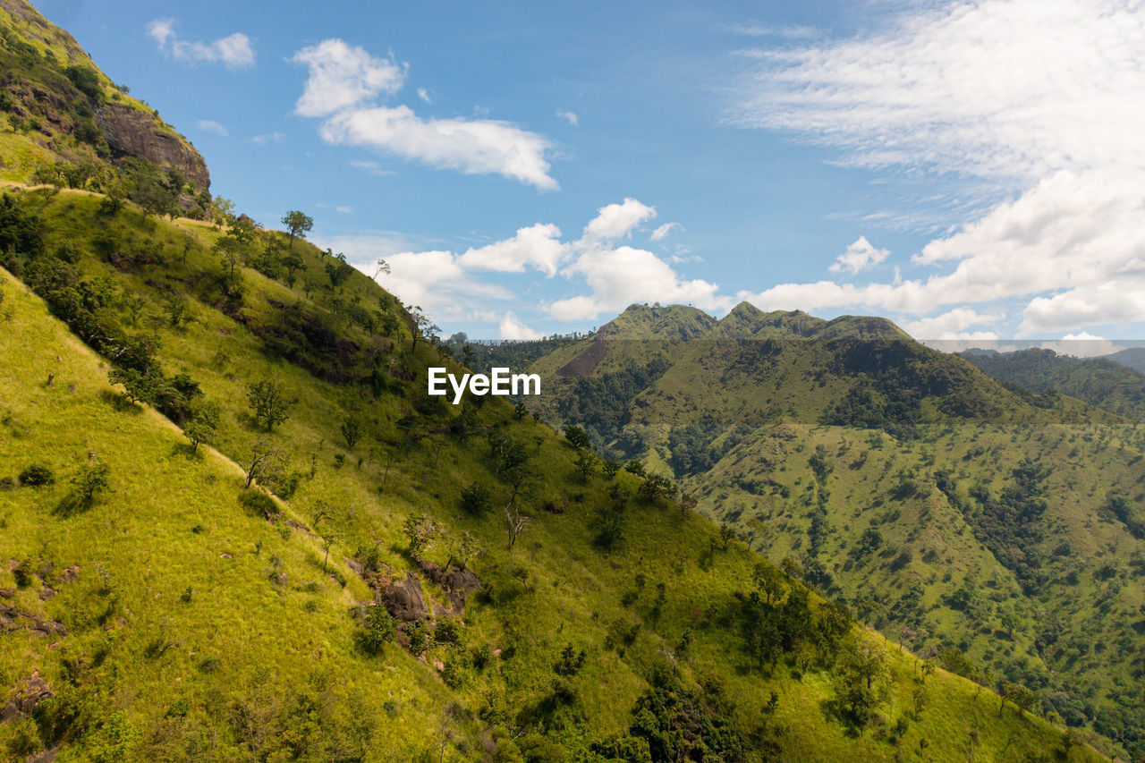 Aerial view of mountain slopes with green forest on the background of blue sky and clouds.