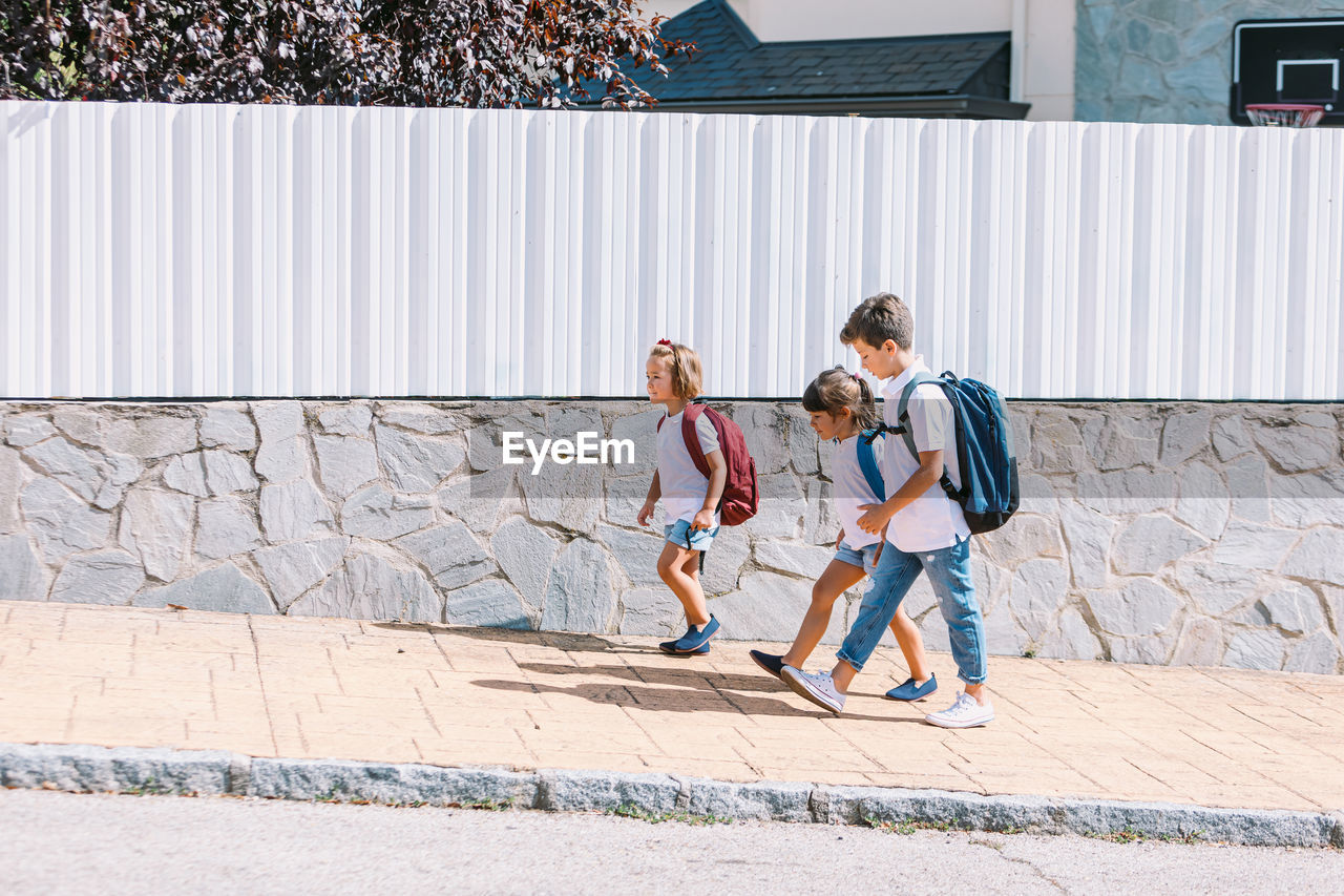 Side view of schoolboy with backpack speaking with female friends while strolling on tiled pavement against stone wall in sunlight