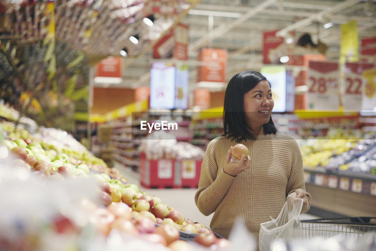 Smiling woman standing in supermarket and holding apple