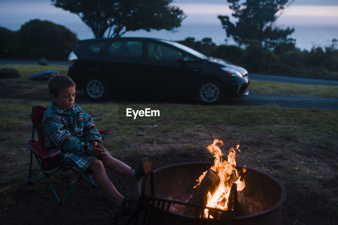 Full length of boy sitting on camping chair by campfire during dusk