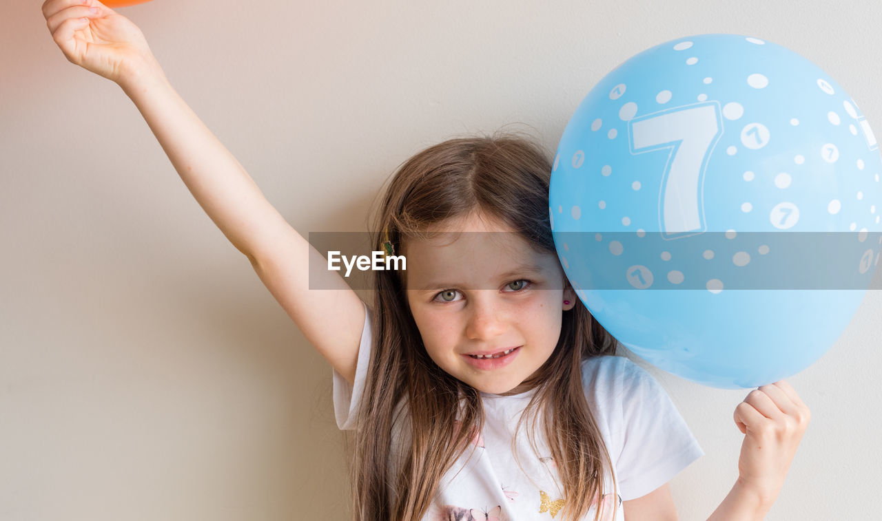 Portrait of cute girl holding blue balloon against wall