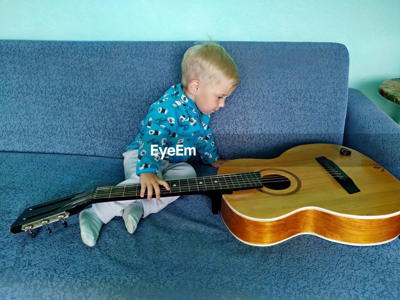 Little boy on a blue couch looks at a retro guitar with interest