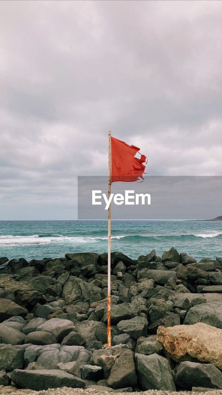 flag, sky, rock, water, cloud, sea, nature, ocean, coast, beach, land, patriotism, shore, red, environment, beauty in nature, no people, horizon, scenics - nature, horizon over water, day, tranquility, wind, outdoors, wave, tranquil scene, red flag