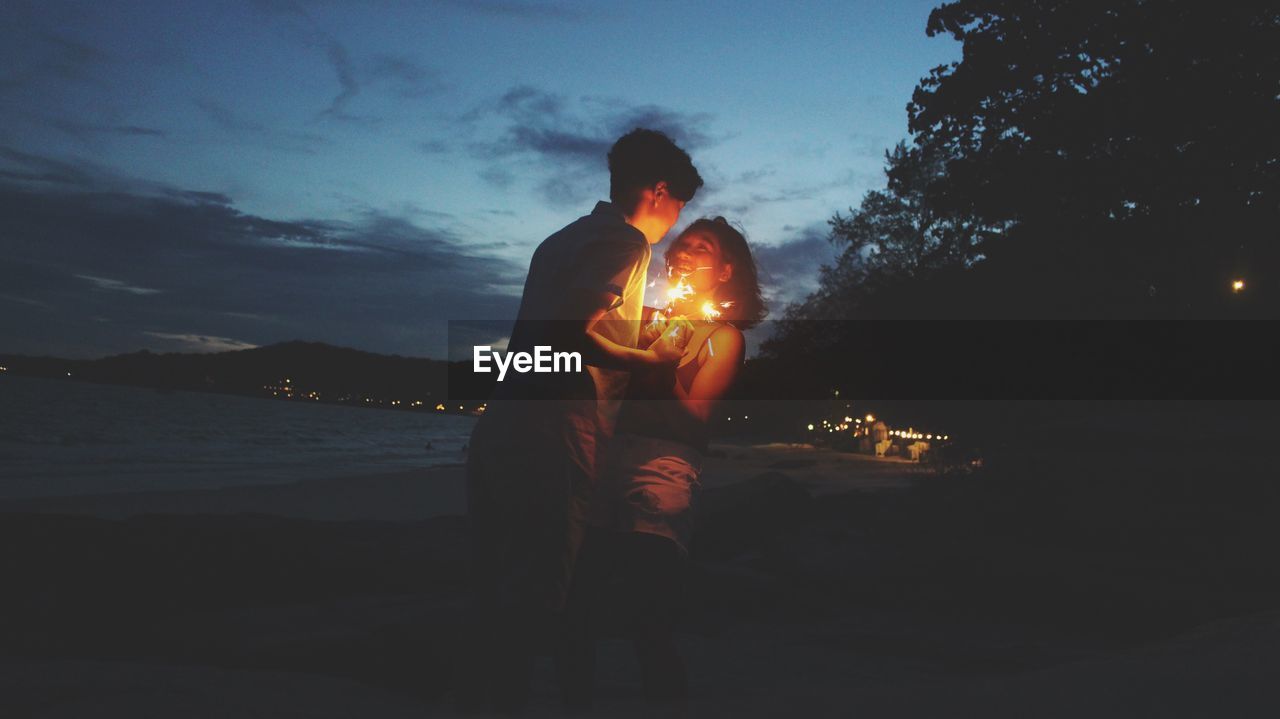 Young couple with illuminated sparklers standing at beach during dusk
