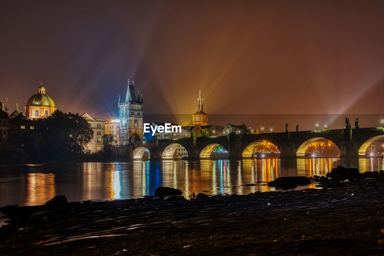 Illuminated bridge over river by buildings against sky at night