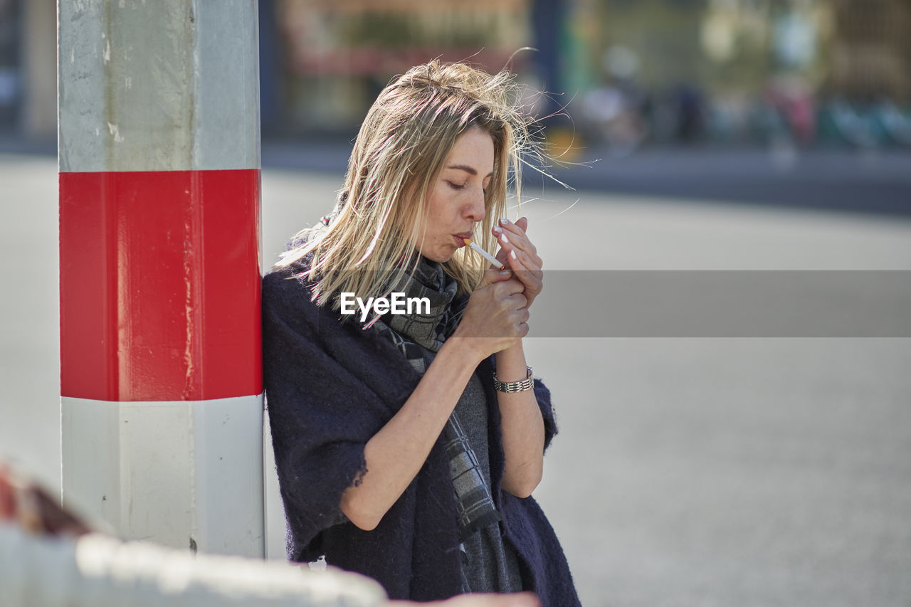 Side view of young female in scarf smoking cigarette near post on urban roadway in back lit