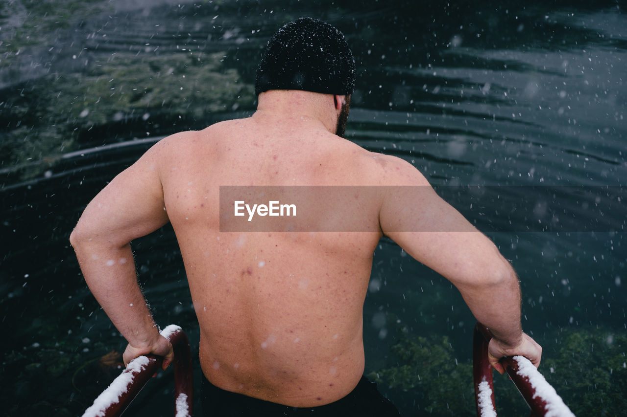 Rear view of shirtless man moving in lake during snowfall in winter