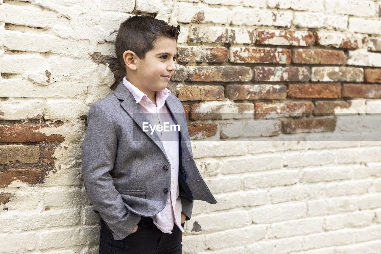 Boy looking away while standing against brick wall