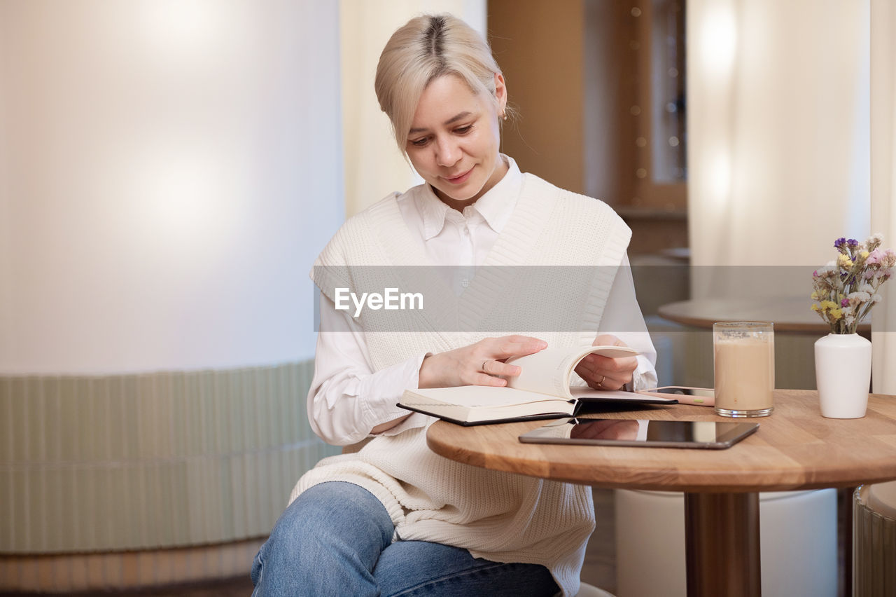 A cute girl in a shirt is flipping through a diary lying next to a tablet and a cup of coffee 