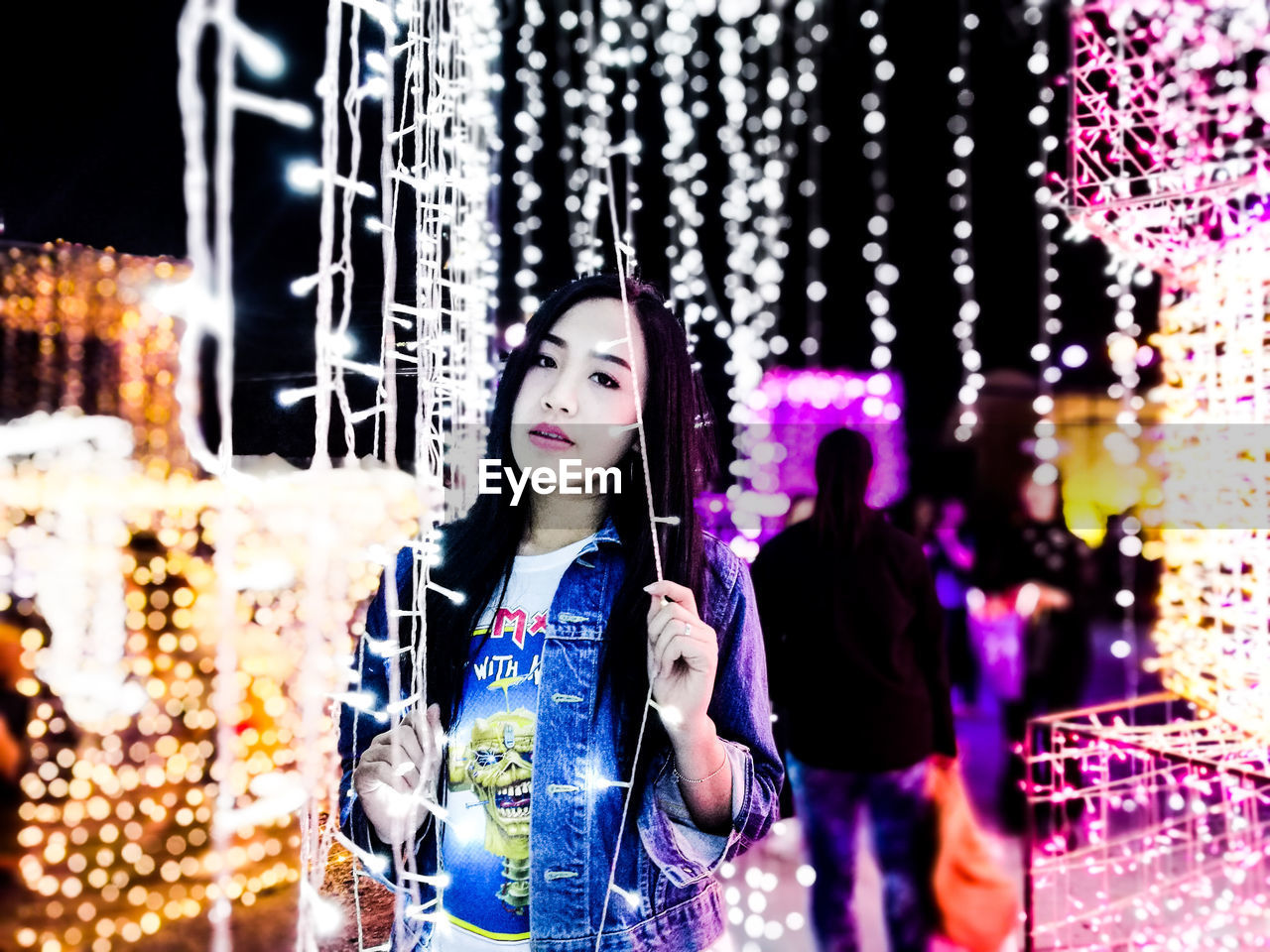 PORTRAIT OF HAPPY YOUNG WOMAN STANDING AGAINST ILLUMINATED WALL