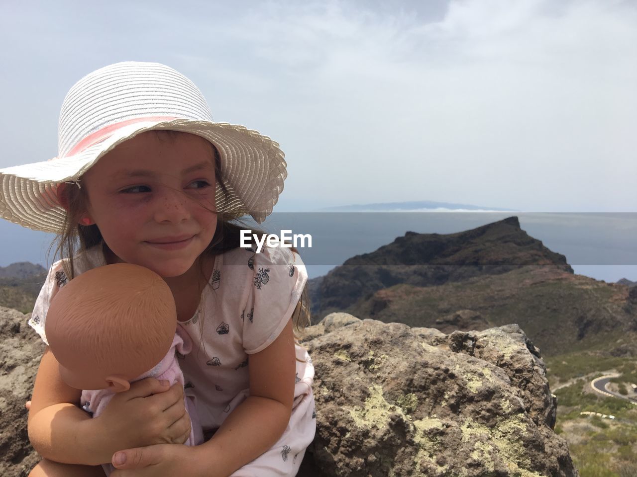 Smiling girl with doll sitting on rock against mountain