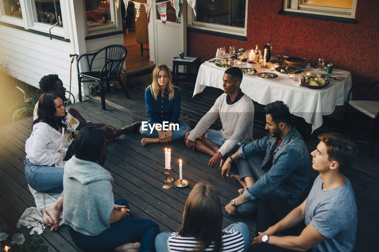 High angle view of male and female friends sitting with eyes closed around lit candles in balcony during group therapy s